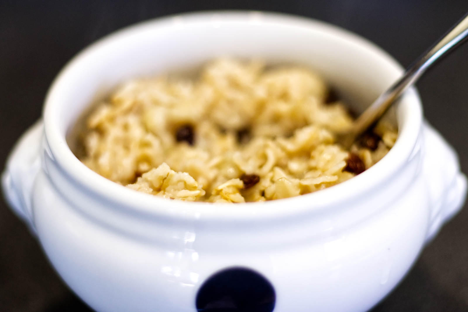 A white bowl with a silver utensil filled with oatmeal, cooked banana, and raisins